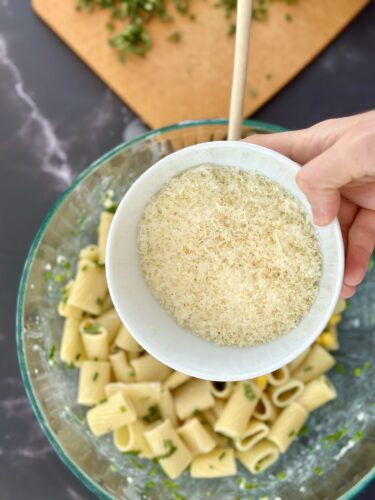 A hand holding a bowl of grated cheese above a glass bowl filled with cooked pasta and chopped herbs.