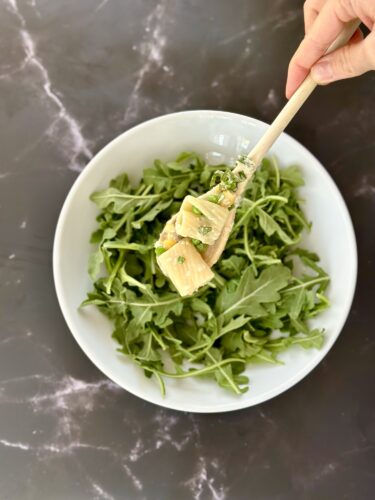 A hand holding a wooden fork with pieces of food above a bowl of fresh arugula on a marble surface.