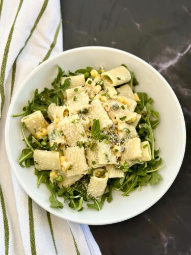 A bowl of rigatoni pasta topped with corn, herbs, and grated cheese, served on a bed of arugula, with a striped kitchen towel nearby.