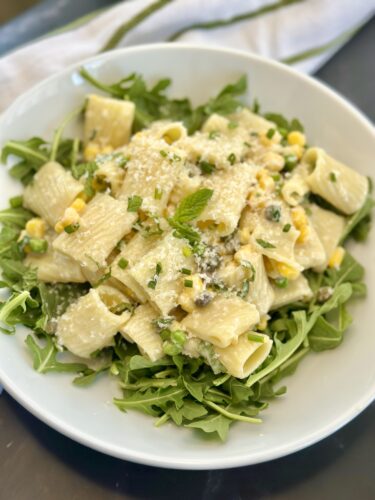 A bowl of rigatoni pasta served on a bed of arugula, topped with corn, herbs, and grated cheese.