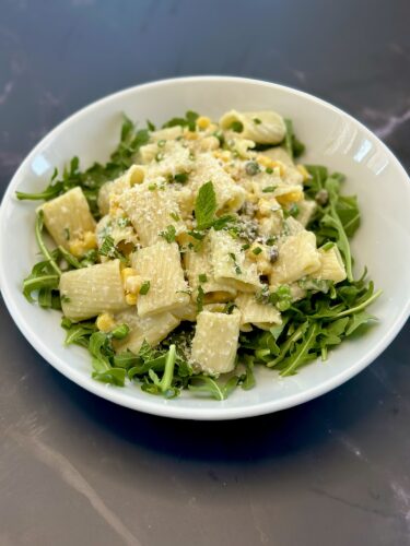 A bowl of rigatoni pasta mixed with corn and peas, served over a bed of fresh arugula and garnished with mint leaves and grated cheese.