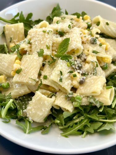A bowl of rigatoni pasta served on a bed of arugula, garnished with mint, chopped green onions, and sprinkled with grated cheese.