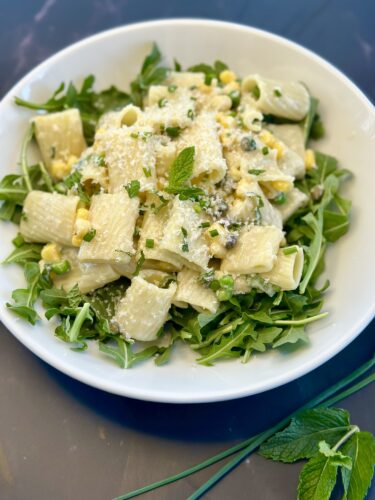 A bowl of rigatoni pasta topped with corn, herbs, and cheese, served on a bed of fresh arugula.