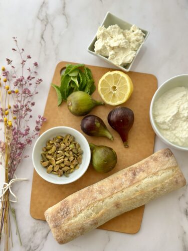 A wooden cutting board displaying various ingredients: a loaf of bread, fresh figs, lemon slices, mint leaves, pistachios, and bowls of ricotta cheese.