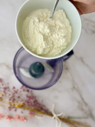 A person holding a bowl of white flour or powdered ingredient above a kitchen food processor, with dried flowers in the foreground.