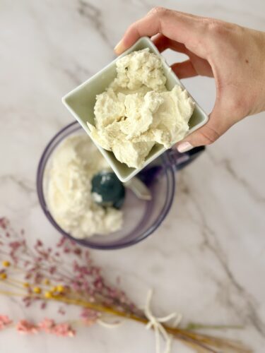A hand holding a small dish of creamy mixture above a food processor containing a similar mixture, with dried flowers in the background.