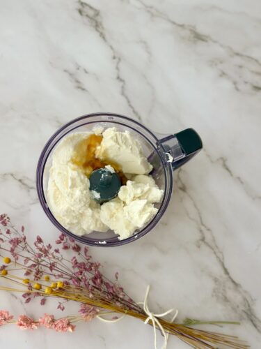 A top view of a food processor bowl containing ricotta cheese, a raw egg, and a splash of vanilla extract, surrounded by dried flowers on a marble countertop.