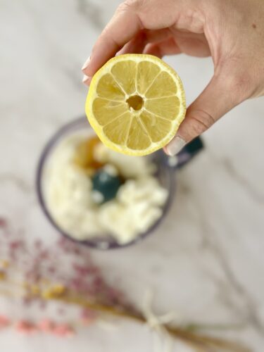 A hand holding a sliced lemon above a bowl containing creamy ingredients, with dried flowers in the background.