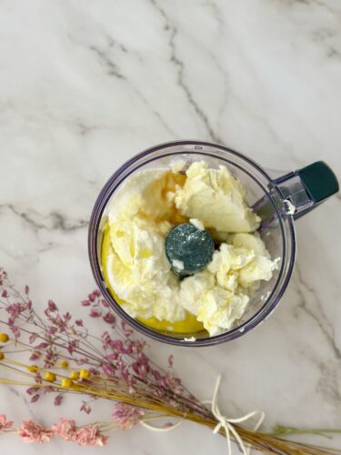A top-down view of a food processor containing a mixture of fluffy white ingredients, likely a blend of cream or cheese, with a dark green processing cap, surrounded by dried flowers on a marble surface.