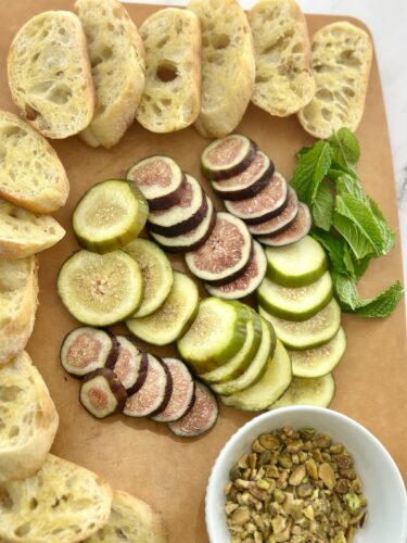 Sliced fresh figs, cucumbers, and mint leaves arranged on a wooden cutting board, accompanied by slices of bread and a bowl of pistachios.