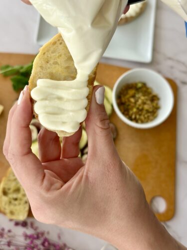 A hand holding a slice of bread with creamy sauce being piped onto it, alongside a bowl of chopped nuts.