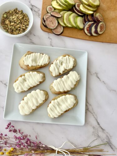 A platter of six iced cookies with a decorative white frosting, placed on a white square plate. In the background, there are sliced figs and a bowl of granola.