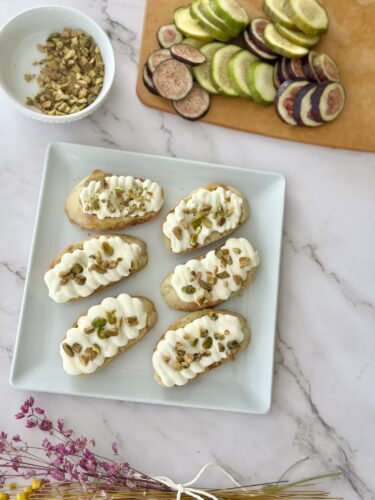 A plate of six dessert canapés topped with whipped cream and chopped pistachios, alongside a wooden cutting board with sliced figs and colorful fruit.