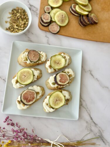 A white plate with six open-faced sandwiches topped with cream cheese, figs, and granola, placed on a marble surface. In the background, sliced figs and a bowl of granola are seen on a wooden board.
