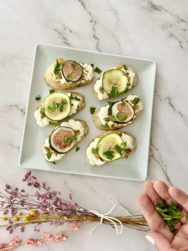 A plate of five decorative crostinis topped with cream cheese, sliced figs, cucumber, and fresh herbs, accompanied by a hand holding fresh herbs in the foreground.