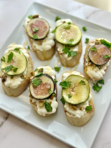 A plate of six open-faced bread appetizers topped with creamy cheese, fresh mint, sliced figs, and crushed nuts.