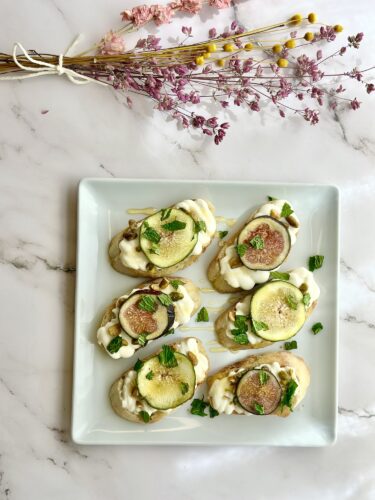 A plate of six fig and cream cheese crostinis, garnished with fresh mint leaves, on a marble surface with dried flowers in the background.