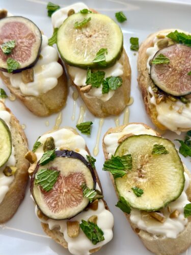A platter of crostini topped with whipped cream, fresh fig slices, crushed nuts, and mint leaves, drizzled with honey.