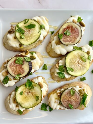 A platter of six open-faced sandwiches topped with cream cheese, figs, mint leaves, and pistachios drizzled with honey.