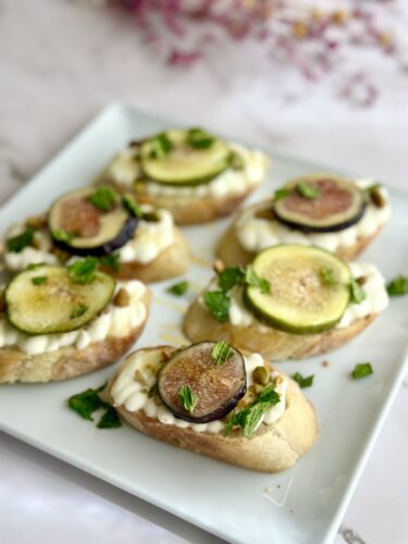 A plate of five bruschetta topped with cream cheese, slices of fresh figs, and zucchini, garnished with mint leaves.