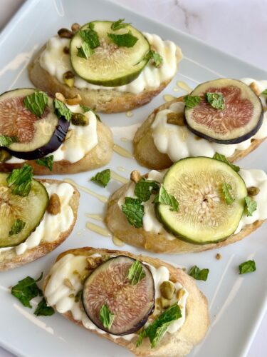 A plate of toasted bread topped with creamy yogurt, fresh figs, sliced cucumbers, pistachios, and mint leaves, drizzled with honey.