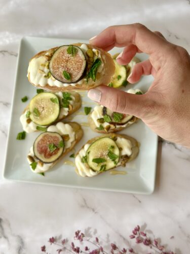 A hand holding a sliced fig topped crostini, with a plate of similar crostini in the background, garnished with mint and yogurt.