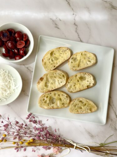 A plate of sliced bread arranged in a pattern, surrounded by bowls of olives and shredded cheese, with decorative flowers in the foreground.