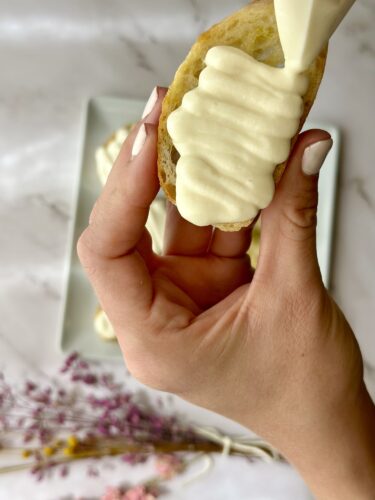 Close-up of a hand holding a piece of bread topped with a creamy sauce, with a plate of similarly topped bread in the background.