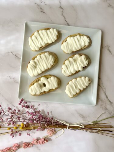 A square white plate with six decorated pastries topped with white frosting, surrounded by dried flowers.