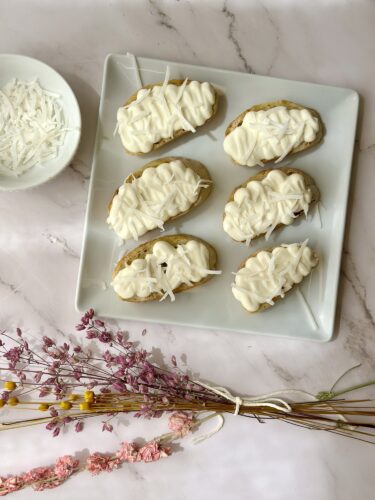 A plate of six decorated cookies with white icing on a marble surface, accompanied by a small bowl of shredded coconut and dried flowers.