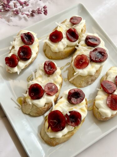 A plate of bruschetta topped with cream cheese, sliced cherries, and drizzled with honey. The presentation is elegant, with a light background and decorative flowers in the corner.