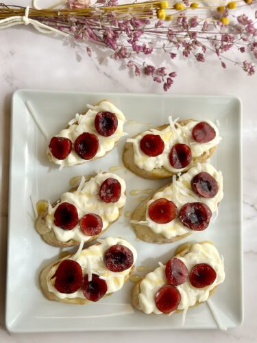 A plate of sliced bananas topped with whipped cream and red fruit jelly, garnished with shredded coconut and surrounded by small purple flowers.