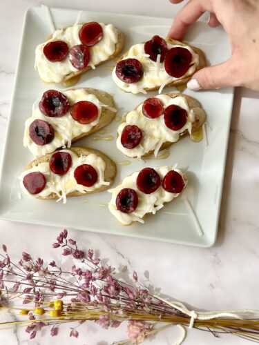 A plate of mini pastries topped with whipped cream, cherries, and a drizzle of honey, with a hand reaching for one of the pastries. A bouquet of dried flowers is placed nearby.