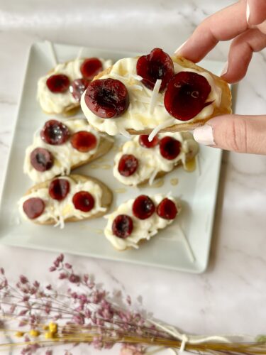 A hand holding a slice of dessert topped with cream and cherries, with additional similar slices arranged on a square plate in the background.