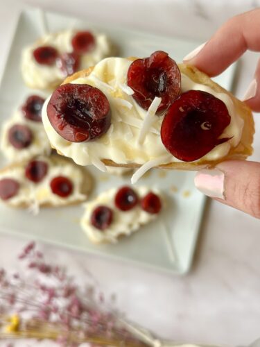 A hand holding a slice of dessert topped with cream and cherries, with more slices in the background on a plate.
