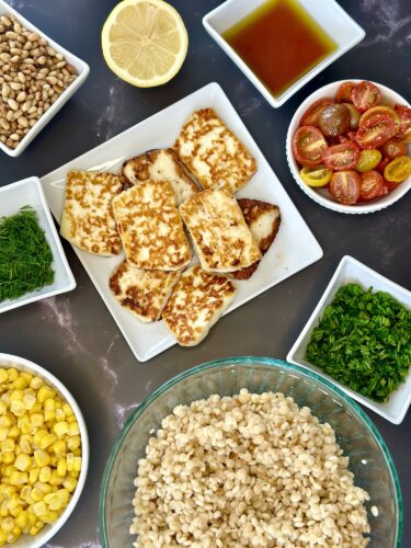 A variety of ingredients arranged on a table, including grilled cheese slices on a white plate, fresh herbs, cherry tomatoes, corn, and bowls of olive oil and lemon, creating a colorful meal preparation scene.