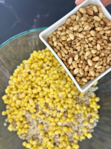 A hand holding a small white dish filled with pine nuts above a large glass bowl containing yellow corn and grains.