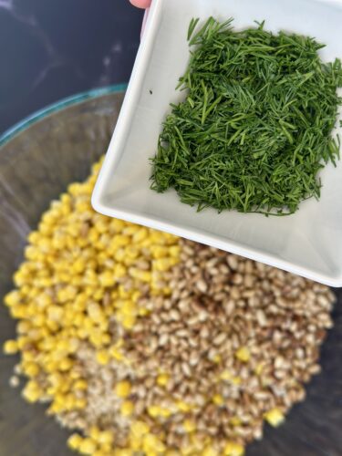 A close-up image showing a bowl containing yellow corn and brown grains, with a small plate holding freshly chopped green herbs, likely dill, above the bowl.
