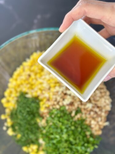 A hand holding a small square dish of brown liquid oil above a mixing bowl with ingredients including corn, chopped herbs, and grains.