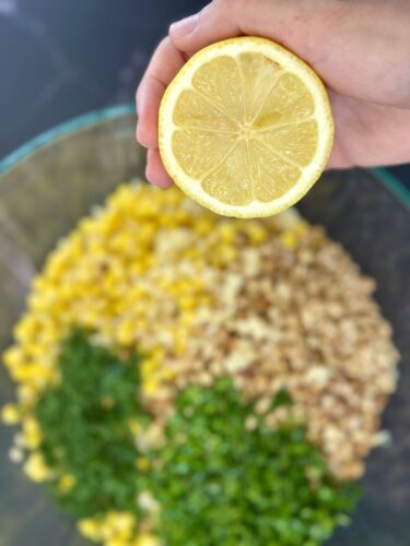 A person holding a half lemon above a bowl filled with yellow corn, chopped herbs, and grains.
