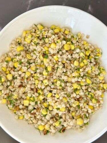 A bowl of cooked pearl couscous mixed with fresh corn and chopped herbs, garnished with yellow corn pieces.