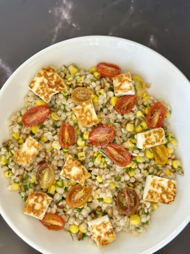 A bowl of pearl couscous salad topped with grilled halloumi cheese, cherry tomatoes, and corn, garnished with fresh herbs.