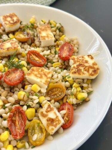 A close-up view of a bowl filled with pearl couscous mixed with grilled halloumi cheese, colorful cherry tomatoes, fresh corn, and herbs.