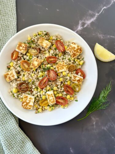 A bowl of barley salad topped with grilled halloumi cheese, cherry tomatoes, and corn, garnished with fresh herbs and a lemon wedge on the side.