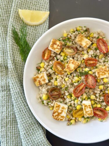 A bowl of pearl couscous salad with grilled tomatoes, corn, and cubed feta cheese, garnished with fresh dill and accompanied by a lemon wedge, set against a textured green cloth.