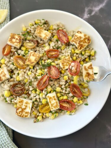 A white bowl filled with barley, roasted cherry tomatoes, corn, and cubes of grilled cheese, garnished with fresh herbs.