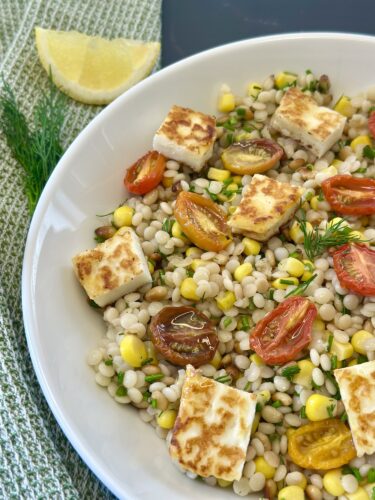 A bowl of pearl couscous salad topped with grilled halloumi cheese, cherry tomatoes, corn, and fresh dill, with a wedge of lemon on the side.