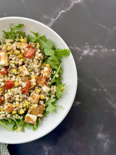 A bowl of salad featuring arugula, cooked barley, roasted cherry tomatoes, and feta cheese on a dark marble surface.