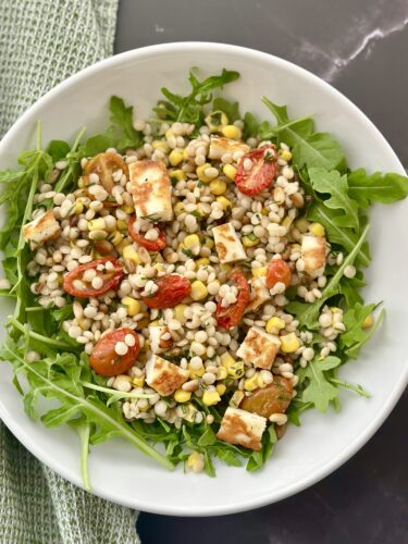 A white bowl filled with a salad made of arugula, cooked grains, cherry tomatoes, corn, and cubes of feta cheese.
