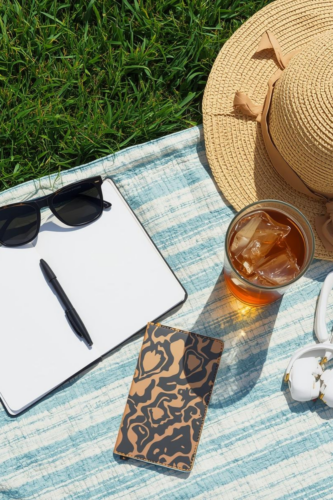 An overhead view of a picnic setup featuring a straw hat, sunglasses, a notebook with a pen, a patterned phone case, and a glass of iced tea on a blue and white striped blanket laid on grass.